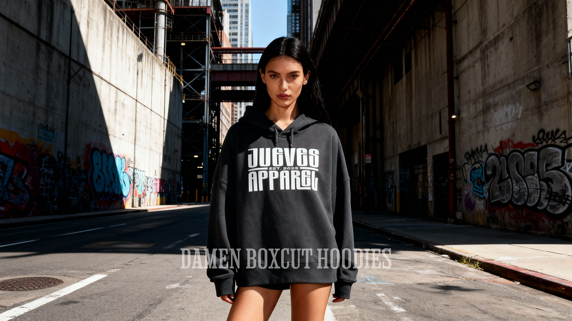 Frau steht auf der Straße und trägt den Damen Boxcut Hoodie mit dem "Long Label" Design auf der Brust in Black. Der Hoodie besteht auf 100% Baumwolle. Moderner Urban Style von Jueves Apparel.