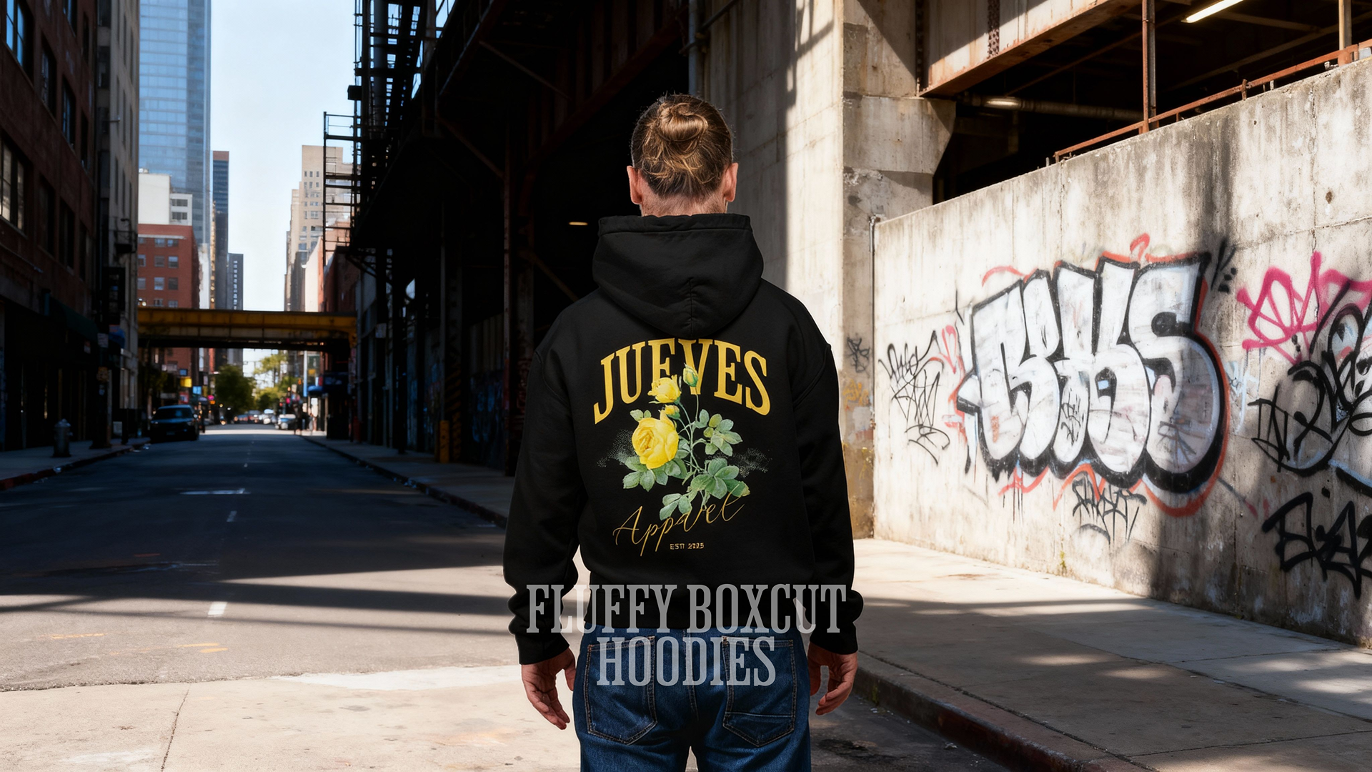 Mann steht auf der Straße und trägt den Fluffy Boxcut Hoodie mit dem "Yellow Rose" Design auf dem Rücken in Black. Der Hoodie besteht aus 30% Polyester und 70% Baumwolle. Moderner Urban Style von Jueves Apparel.
