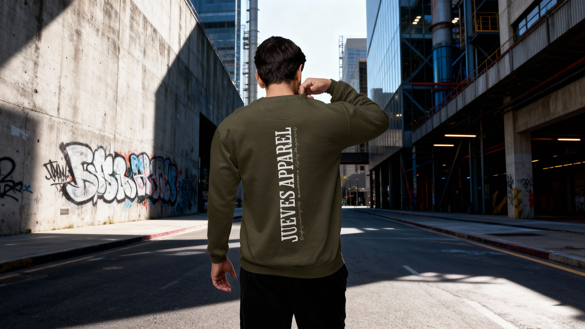 Mann steht auf der Straße und trägt das Oversized Sweatshirt mit dem "All you need" Design auf dem Rücken in Khaki. Das Sweatshirt besteht aus 100% Baumwolle. Moderner Urban Style von Jueves Apparel.